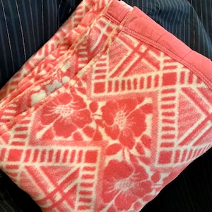 May include: Folded coral-colored blanket with a floral and geometric pattern. The blanket features a repeating design of flowers and geometric shapes in white against a coral background. The edge is trimmed with a coral border.