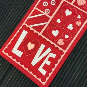 May include: A red Valentine's Day table runner with the word "LOVE" in white, polka-dotted letters. The runner features various heart designs and geometric patterns in red and white, with a scalloped edge.