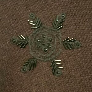 May include: A brown knitted fabric with a decorative snowflake design made of silver beads. The snowflake has six points and a small circle in the center.