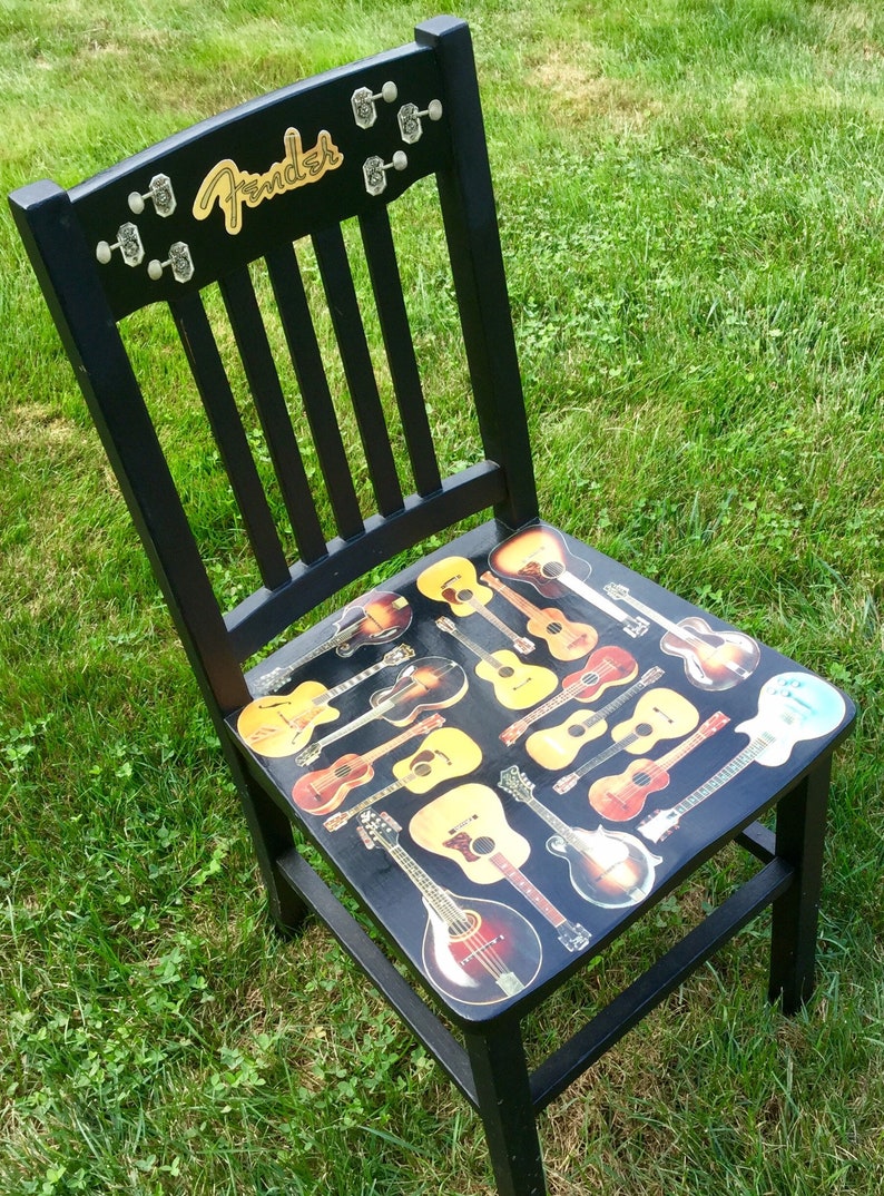 Op de afbeelding: Een zwarte houten stoel met een Fender-logo op de rugleuning. De zitting is bedekt met een collage van verschillende gitaar afbeeldingen.