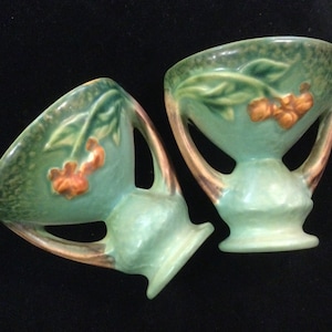 May include: Two green glazed ceramic vases with brown handles and floral designs. The vases have a textured surface and are decorated with orange flowers and green leaves.