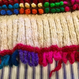 K&ouml;nnte beinhalten: Nahaufnahme eines gestrickten Stoffes mit einem Fransenrand. Die Fransen bestehen aus buntem Garn in den Farben Rot, Rosa, Lila, Blau, Gr&uuml;n und Wei&szlig;. Der Stoff ist cremefarben und hat ein strukturiertes Strickmuster.