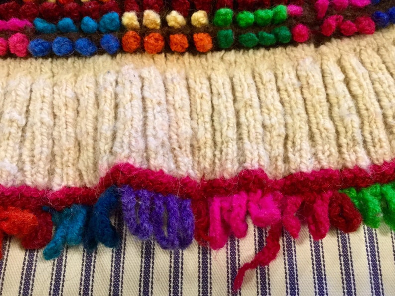 K&ouml;nnte beinhalten: Nahaufnahme eines gestrickten Stoffes mit einem Fransenrand. Die Fransen bestehen aus buntem Garn in den Farben Rot, Rosa, Lila, Blau, Gr&uuml;n und Wei&szlig;. Der Stoff ist cremefarben und hat ein strukturiertes Strickmuster.