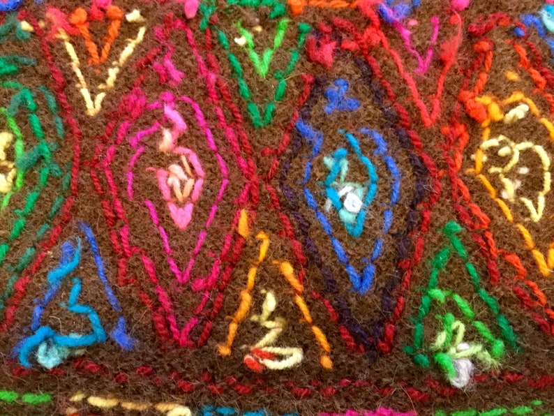 K&ouml;nnte beinhalten: Nahaufnahme eines braunen Stoffes mit bunter Stickerei. Die Stickerei zeigt ein sich wiederholendes Muster aus Rauten und Dreiecken in Rot-, Rosa-, Blau-, Gr&uuml;n- und Gelbt&ouml;nen.