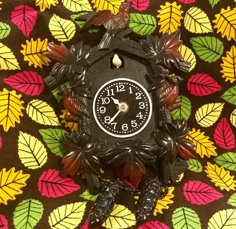 Op de afbeelding: Een zwarte koekoeksklok met een witte wijzerplaat en zwarte wijzers. De klok is versierd met gesneden houten bladeren en vogels. De klok hangt aan een ketting met twee zwarte kwastjes.