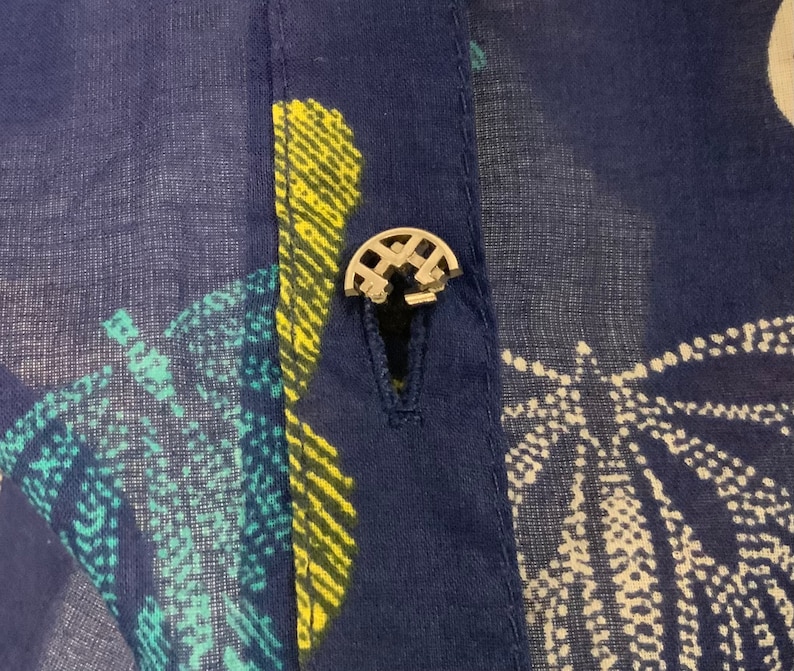 May include: Close-up of a navy blue fabric with a buttoned closure. The fabric has a pattern of white, yellow, and teal shapes. The button is silver and has a decorative design.