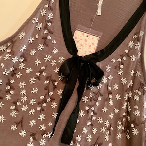 May include: A gray sleeveless top with a black bow and white floral embroidery. The top has a scoop neck and a relaxed fit.