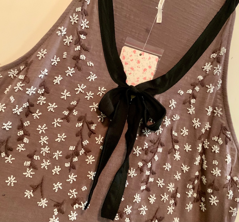 May include: A gray sleeveless top with a black bow and white floral embroidery. The top has a scoop neck and a relaxed fit.