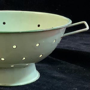 May include: A vintage, light green colander with a sturdy handle. The colander features a series of evenly spaced holes for draining liquids. The rim and handle are trimmed with a darker green. This kitchenware item is ideal for straining pasta and vegetables.