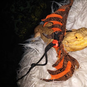 May include: A bearded dragon wearing a brown and orange dragon costume with black straps and a red clip. The dragon is sitting on a white fluffy surface.