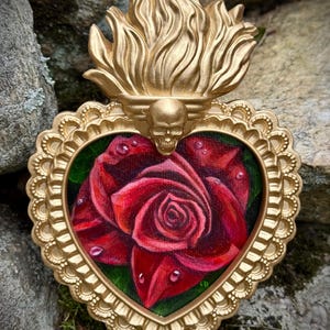 Peut inclure: Un ornement mural en forme de cœur présentant une peinture centrale d'une rose rouge foncé avec des gouttelettes d'eau. L'œuvre est encadrée par un cadre doré orné, avec un crâne sculpté surmonté d'un motif de flamme dorée stylisée.