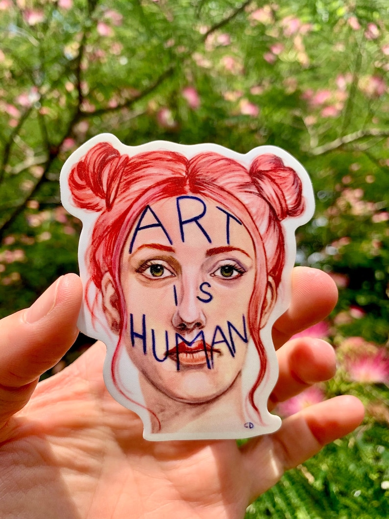 May include: A white sticker with a drawing of a woman with red hair in two buns. The woman's face is covered with the words "ART IS HUMAN" in blue.