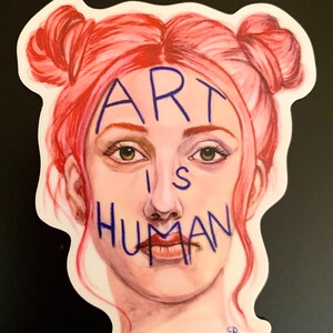 May include: A sticker with a drawing of a person with pink hair in two buns. The person's face is covered with blue writing that says "ART IS HUMAN".
