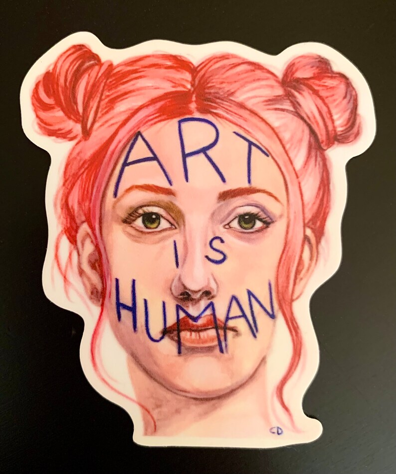 May include: A sticker with a drawing of a person with pink hair in two buns. The person's face is covered with blue writing that says "ART IS HUMAN".