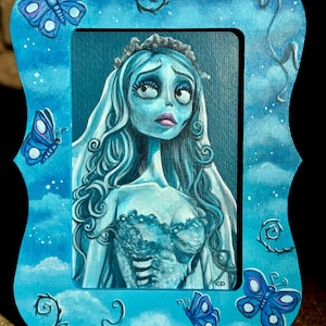 May include: A hand-painted wooden frame with a blue background and white clouds. The frame features a blue and white butterfly design and a portrait of the character Sally from the movie "The Nightmare Before Christmas".