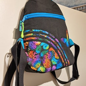 May include: A black backpack with a blue zipper and a colourful butterfly print. The backpack has adjustable straps and a small pocket on the front.