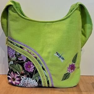 May include: A green corduroy tote bag with a floral print on the bottom and a dragonfly applique. The bag has a purple and gold trim.