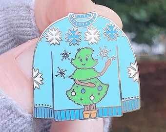 Può includere: Spilla smaltata azzurra a forma di maglione natalizio. Il maglione presenta un albero di Natale verde con un volto sorridente, fiocchi di neve e un bordo dorato. La spilla è tenuta in mano. La spilla è di Becka Higham Illustrations.