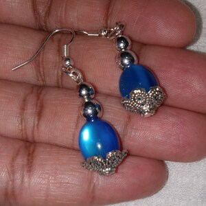 May include: A pair of dangle earrings featuring blue oval-shaped beads with silver accents. The beads have a shimmering finish and are surrounded by a silver filigree design.