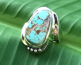 Puede incluir: Anillo de plata con una gran piedra turquesa ovalada con patrones marrones. El anillo tiene una banda de plata pulida y un engaste decorativo con pequeños acentos redondos. La turquesa es de un color azul-verdoso vibrante.