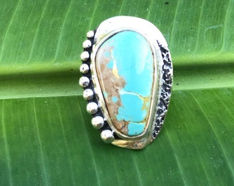 Puede incluir: Un anillo de plata con una piedra turquesa engastada en un engaste texturizado. El anillo tiene un borde con cuentas y una banda texturizada.