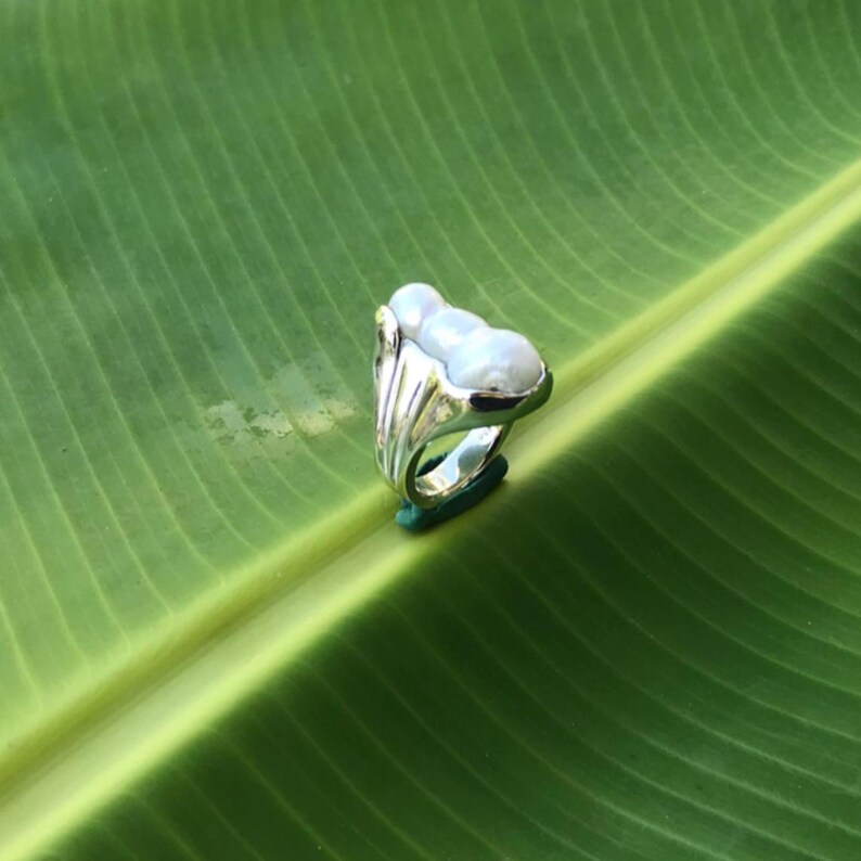 K&ouml;nnte beinhalten: Ein silberner Ring mit drei wei&szlig;en, oval geformten Perlen. Der Ring hat ein einzigartiges Design mit einem strukturierten Band und einer dekorativen Fassung f&uuml;r die Perlen. Der Ring wird auf einer gr&uuml;nen Oberfl&auml;che vor einem gr&uuml;nen Blatt-Hintergrund pr&auml;sentiert.