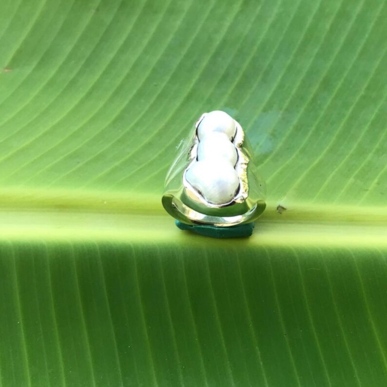 K&ouml;nnte beinhalten: Ein Silberring mit drei wei&szlig;en Perlen. Der Ring liegt auf einer gr&uuml;nen Oberfl&auml;che vor einem gr&uuml;nen Blatt-Hintergrund. Die Perlen sind der Blickfang, mit dem Silberband, das sich um sie legt.