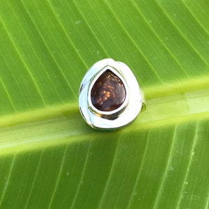 May include: A silver ring with a teardrop-shaped brown gemstone set in a silver bezel. The ring is on a green leaf background.