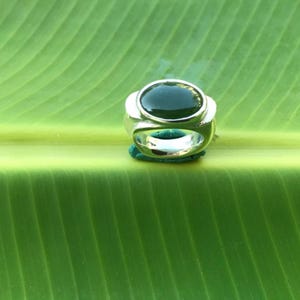 May include: A silver ring with a large, oval, dark green stone. The ring is set on a bright green leaf, showcasing the contrast between the metal and the natural background. The ring's design is simple yet elegant.