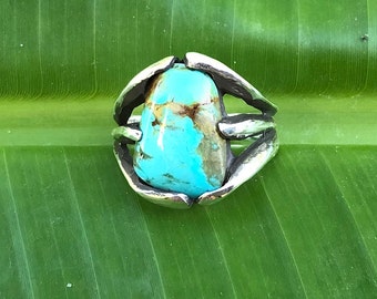Puede incluir: Un anillo de plata con una piedra turquesa engastada en un diseño único y orgánico. El anillo está sobre un fondo de hoja verde.
