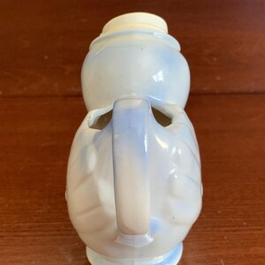 May include: A white ceramic pitcher with a blue glaze and a handle. The pitcher is shaped like a bird with a round body and a long neck.