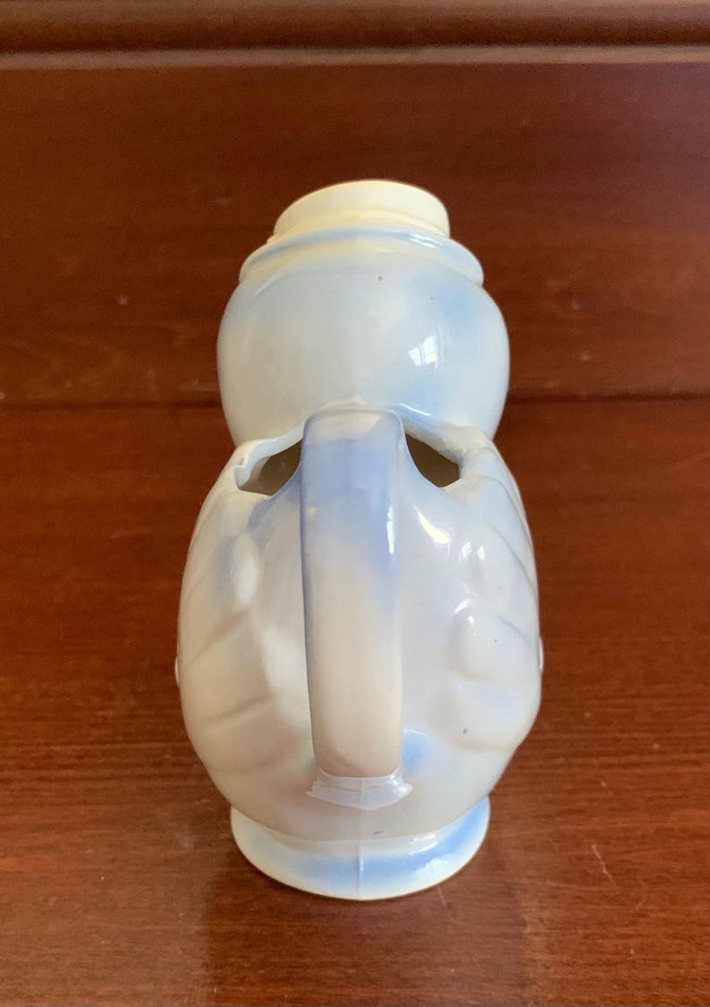 May include: A white ceramic pitcher with a blue glaze and a handle. The pitcher is shaped like a bird with a round body and a long neck.