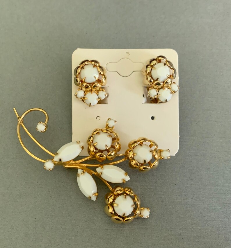 May include: A vintage jewelry set featuring a gold-tone brooch and matching clip-on earrings. The brooch is designed as a floral spray with white cabochon accents. The earrings and brooch have a gold-tone metal setting.