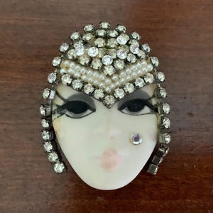 May include: A brooch shaped like a face with a pale complexion, featuring dark painted eyes, pink lips, and a single rhinestone beauty mark. The head is framed by a border of clear rhinestones and a V-shaped band of small white seed pearls.