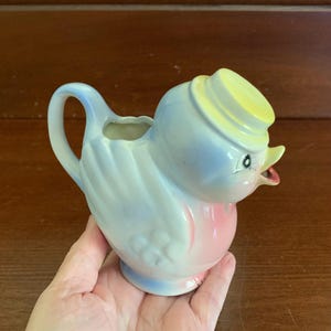 May include: A blue and pink ceramic bird creamer with a yellow hat. The bird has a small spout and a handle.
