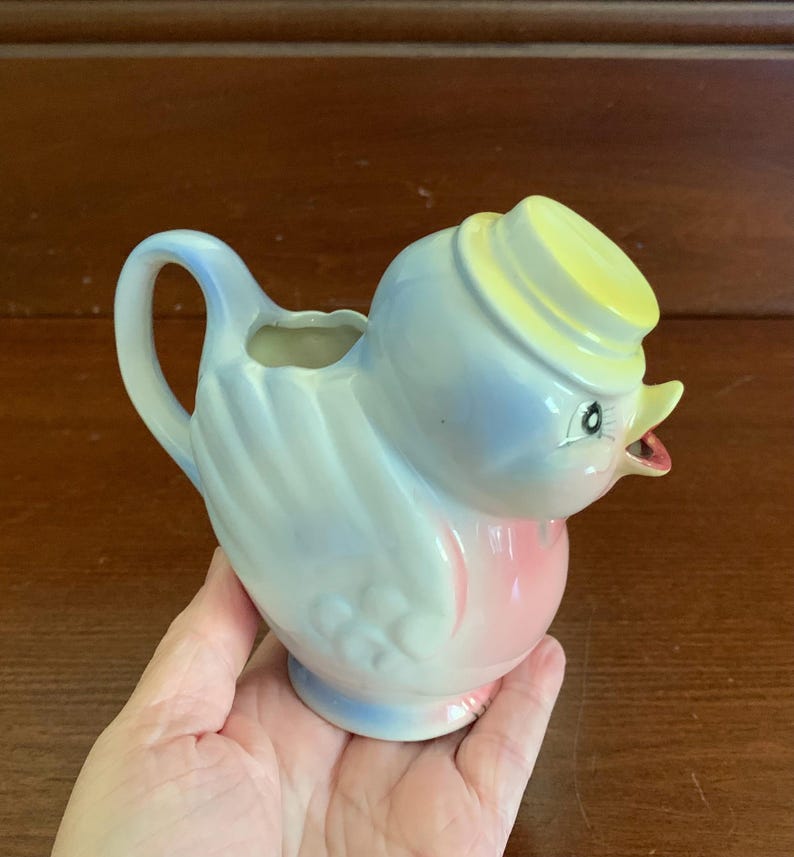May include: A blue and pink ceramic bird creamer with a yellow hat. The bird has a small spout and a handle.