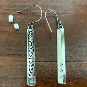Puede incluir: Pendientes plateados con dise&ntilde;o rectangular. Uno presenta un patr&oacute;n de remolino grabado, el otro tiene tres peque&ntilde;as gemas transparentes. Los pendientes tienen cierre de gancho y se exhiben sobre una superficie de madera.