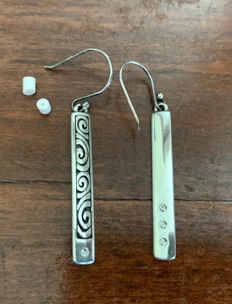 Puede incluir: Pendientes plateados con dise&ntilde;o rectangular. Uno presenta un patr&oacute;n de remolino grabado, el otro tiene tres peque&ntilde;as gemas transparentes. Los pendientes tienen cierre de gancho y se exhiben sobre una superficie de madera.