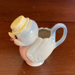 May include: A blue and white ceramic creamer in the shape of a bird wearing a yellow hat. The creamer has a pink belly and a spout on the side.