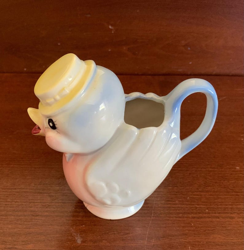 May include: A blue and white ceramic creamer in the shape of a bird wearing a yellow hat. The creamer has a pink belly and a spout on the side.
