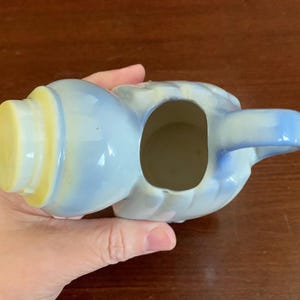May include: A small, blue and white ceramic creamer with a yellow lid and a handle. The creamer is shaped like a person with a rounded body and a small spout.
