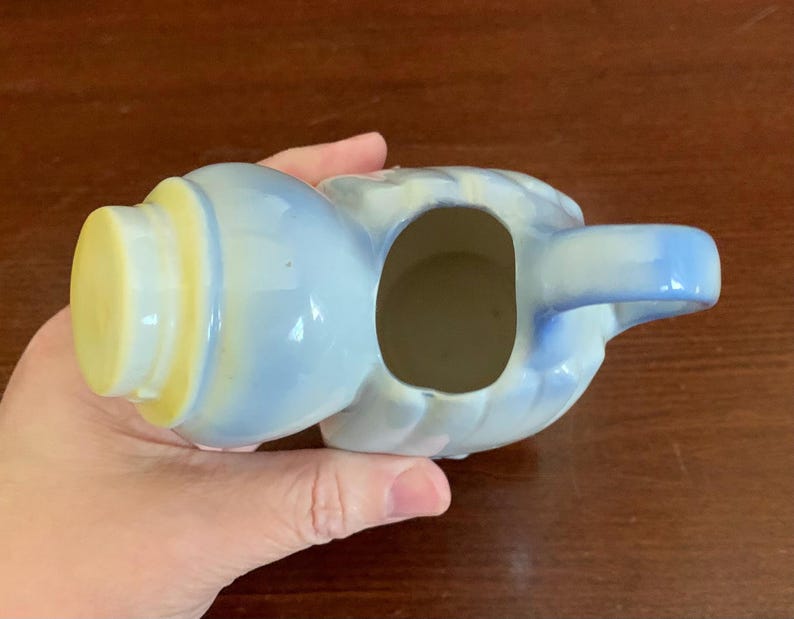 May include: A small, blue and white ceramic creamer with a yellow lid and a handle. The creamer is shaped like a person with a rounded body and a small spout.