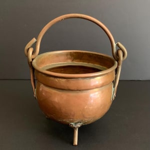 May include: A small, round copper cauldron with a smooth, metallic finish and a warm, reddish-brown hue. The vessel features three short, sturdy legs, a wide open top, and a curved, hinged handle attached to two side loops for easy carrying.