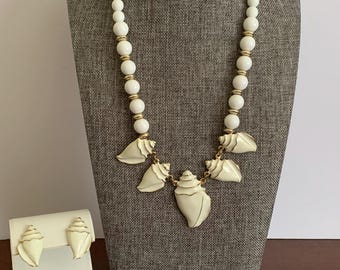 May include: A necklace and matching earring set featuring cream-colored shell-shaped pendants with gold-tone trim. The necklace consists of round white beads interspersed with small gold-tone spacers, leading to five hanging shell charms.