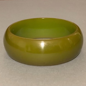 May include: A solid olive green bangle bracelet. The bracelet is a thick, rounded band with a smooth, glossy finish. The color is a muted, earthy green, and the bracelet appears to be made of a solid material.