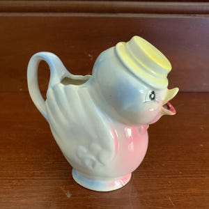 May include: A ceramic creamer in the shape of a blue and pink bird wearing a yellow hat. The bird has a small spout and a handle.