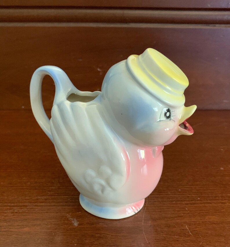 May include: A ceramic creamer in the shape of a blue and pink bird wearing a yellow hat. The bird has a small spout and a handle.