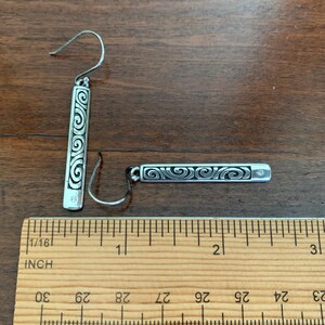 Puede incluir: Un par de pendientes de plata con un dise&ntilde;o rectangular con un patr&oacute;n de remolino. Cada pendiente tiene una peque&ntilde;a gema transparente y mide aproximadamente 2,5 cm de largo.
