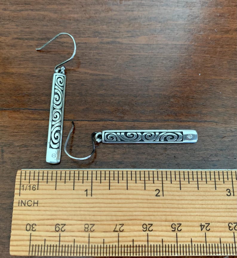 Puede incluir: Un par de pendientes de plata con un dise&ntilde;o rectangular con un patr&oacute;n de remolino. Cada pendiente tiene una peque&ntilde;a gema transparente y mide aproximadamente 2,5 cm de largo.