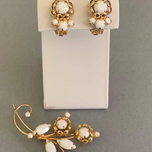 May include: A set of vintage jewelry featuring a pair of clip-on earrings and a brooch. The earrings and brooch have a gold-tone metal setting with white, round, and oval-shaped accents. The earrings are mounted on a white display stand.